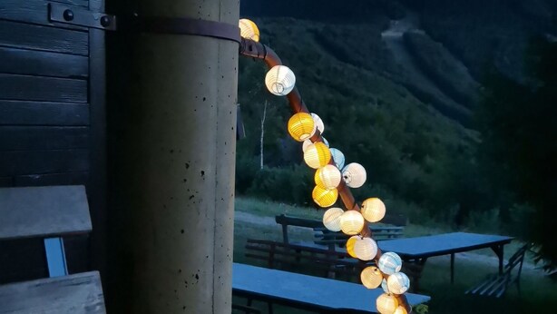 A wooden deck with a table and chairs, a lantern, and string lights against a backdrop of hills. - Le randonneur (Garessio)
