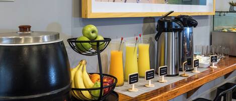 A breakfast bar with a fruit basket, juice dispensers, a coffee pot, and a sign indicating the type of juice available.