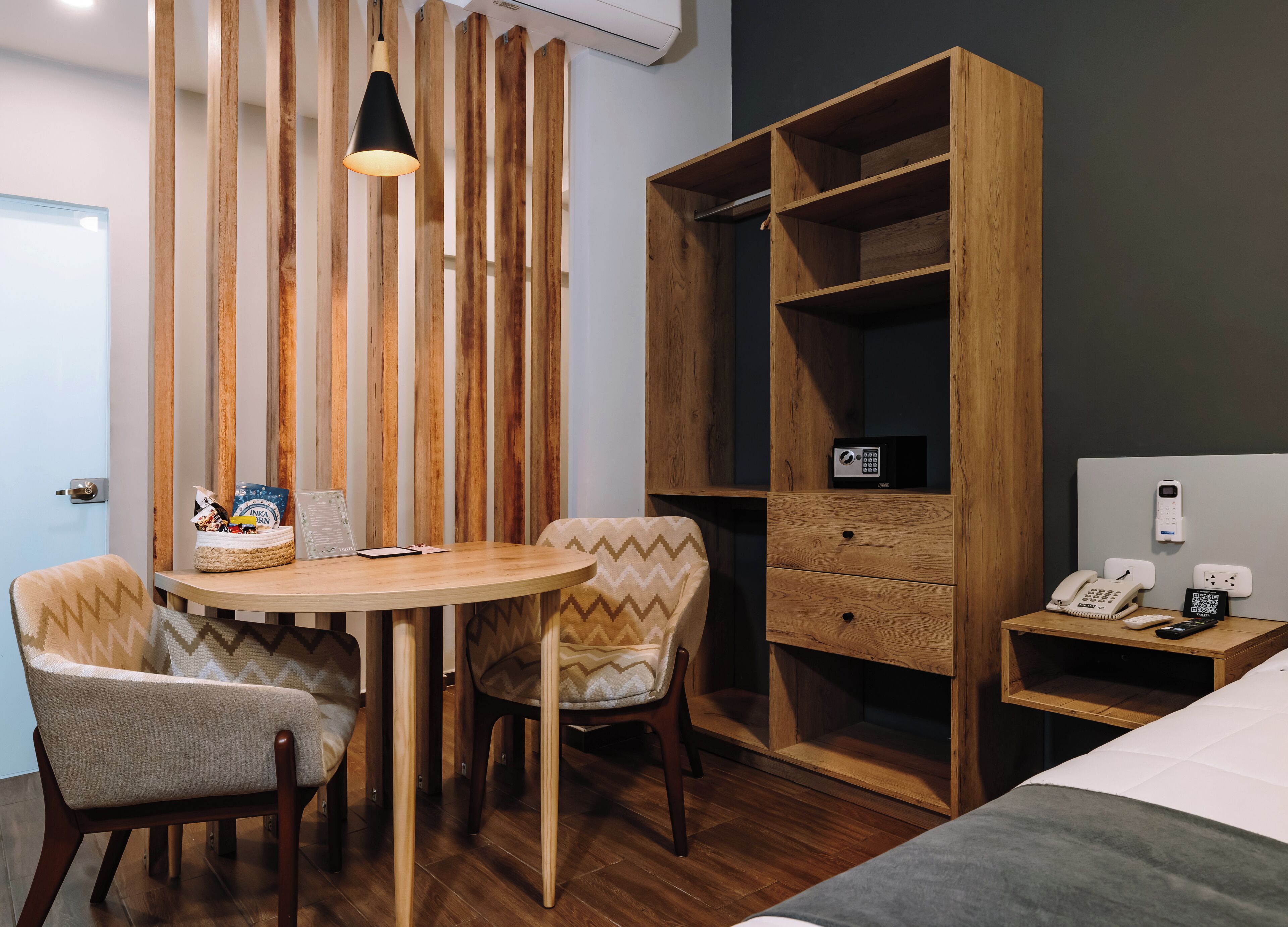 Habitación de hotel con un armario de madera, una mesa redonda con sillas, una mesita de noche con un teléfono y un control remoto, y un aire acondicionado de pared.