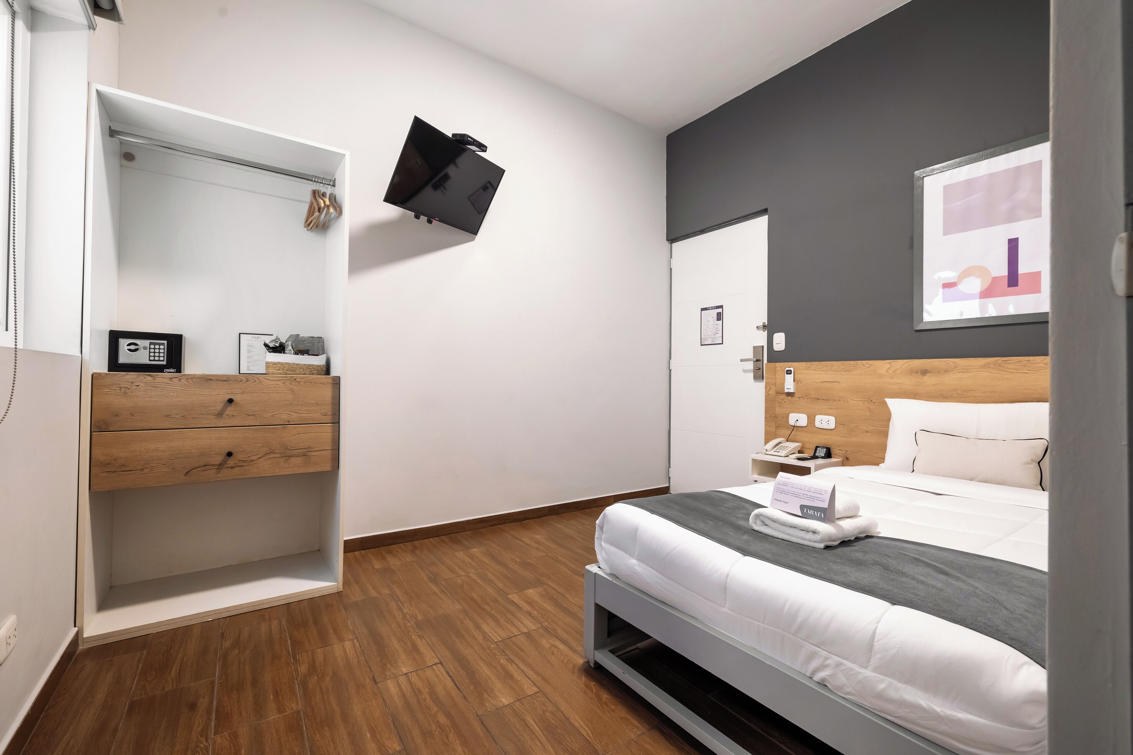 Quarto de hotel moderno com cama, mesa de cabeceira, televisão presa à parede e guarda-roupa de madeira.