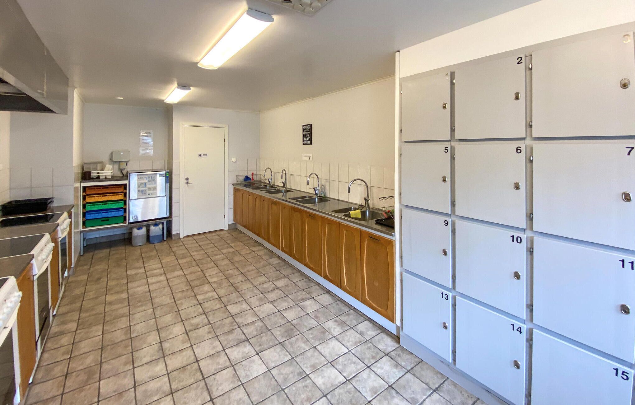 A kitchen with a tiled floor, wooden cabinets, a sink, and a refrigerator. There are storage lockers on the right side.