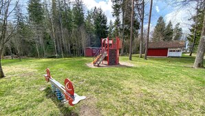 Un parque infantil con un sube y baja, una estructura de escalada y un edificio rojo en un área boscosa.