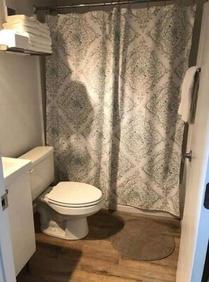 A small bathroom with a toilet, a shower curtain with a pattern, and a wooden floor. - Amazing Beach Studio in Daytona! (Daytona Beach)