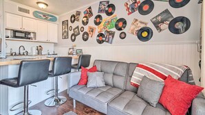 Ein modernes Wohnzimmer mit einer grauen Eckcouch, zwei schwarzen Barhockern und einer Küchenzeile mit weißen Schränken und einer Mikrowelle.