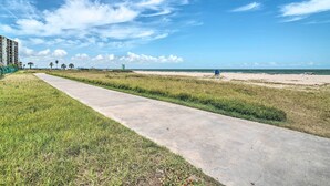 Ein Betonweg führt zu einem Strand mit blauem Himmel und Palmen in der Ferne.