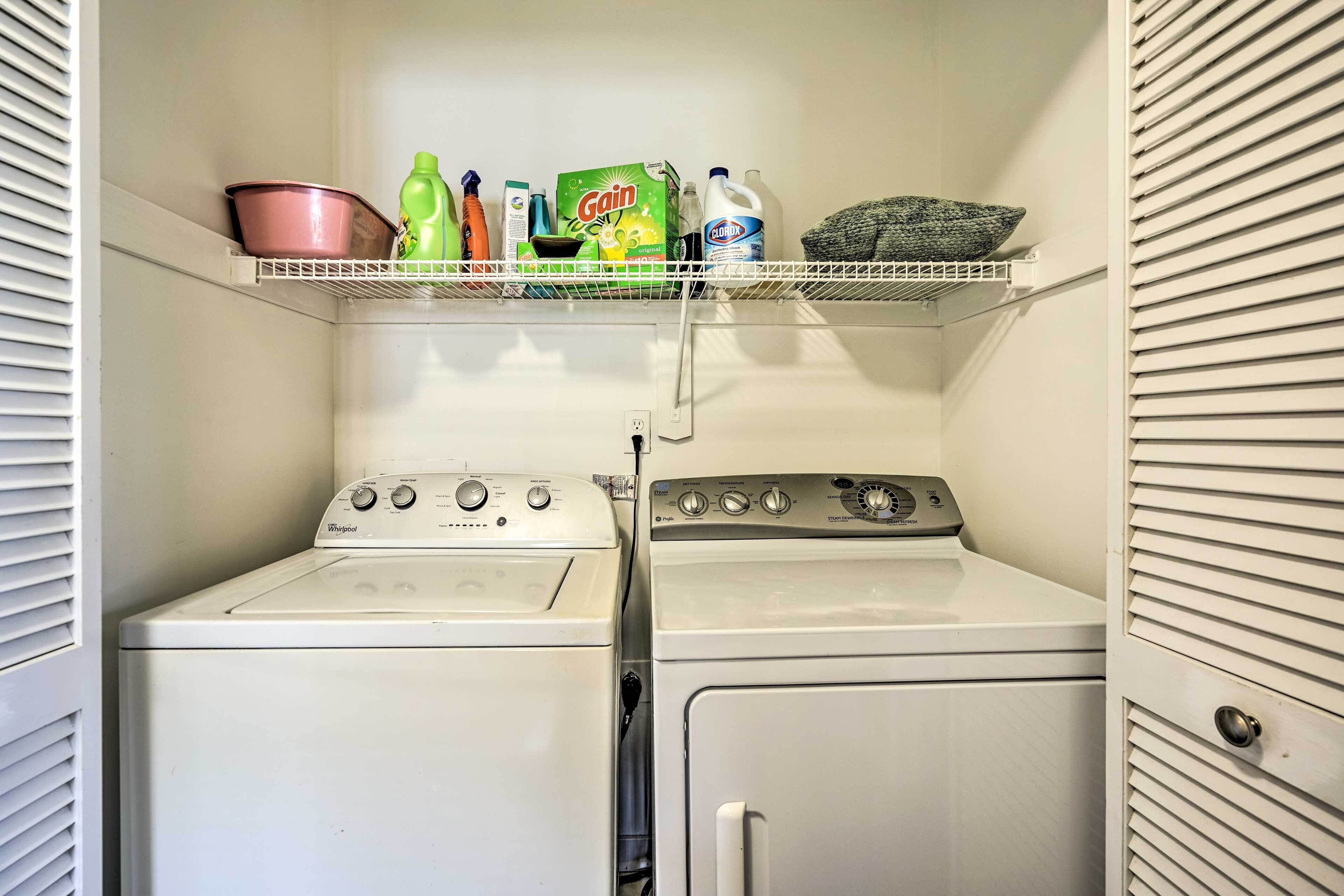 A laundry room with two washers and dryers, shelves above containing cleaning supplies, and a power outlet on the wall.