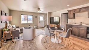 A modern living room with a sofa, a coffee table, and a dining area with chairs. There is a kitchen with dark cabinets and a TV. - Belgrade Vacation Rental w/ Mountain Views! (Belgrade)