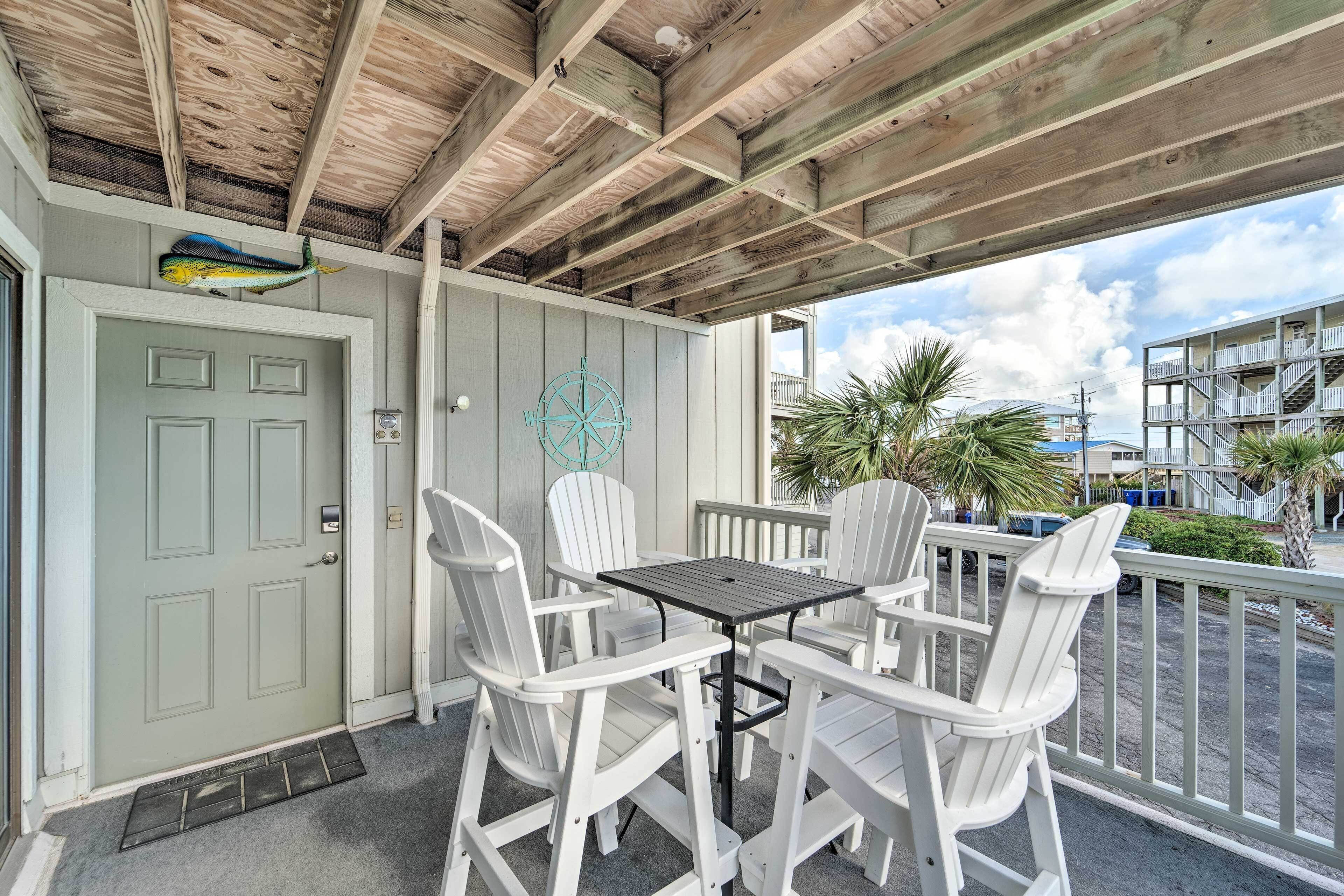 A covered outdoor seating area with white Adirondack chairs and a table, a door with a decorative fish, and a view of a building with balconies.
