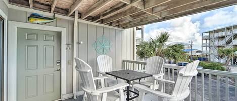 A covered outdoor seating area with white Adirondack chairs and a table, a door with a decorative fish, and a view of a building with balconies.