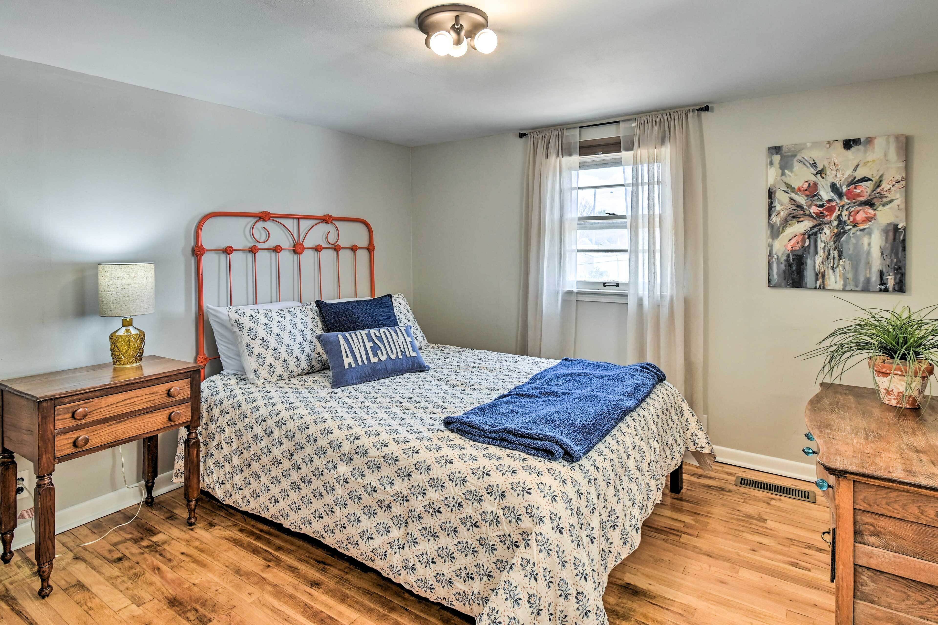 A bedroom with a bed, a nightstand, a lamp, a window with curtains, and a painting on the wall.