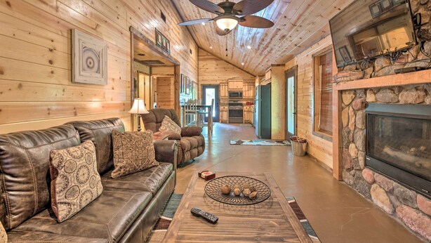 A cozy log cabin interior with a leather sofa, a fireplace, a ceiling fan, and a kitchen in the background. - Cabin w/ Hot Tub - Near Broken Bow Lake & Hiking! (Broken Bow)
