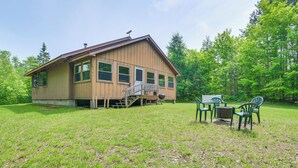 A wooden cabin with a deck, surrounded by a grassy yard and trees.