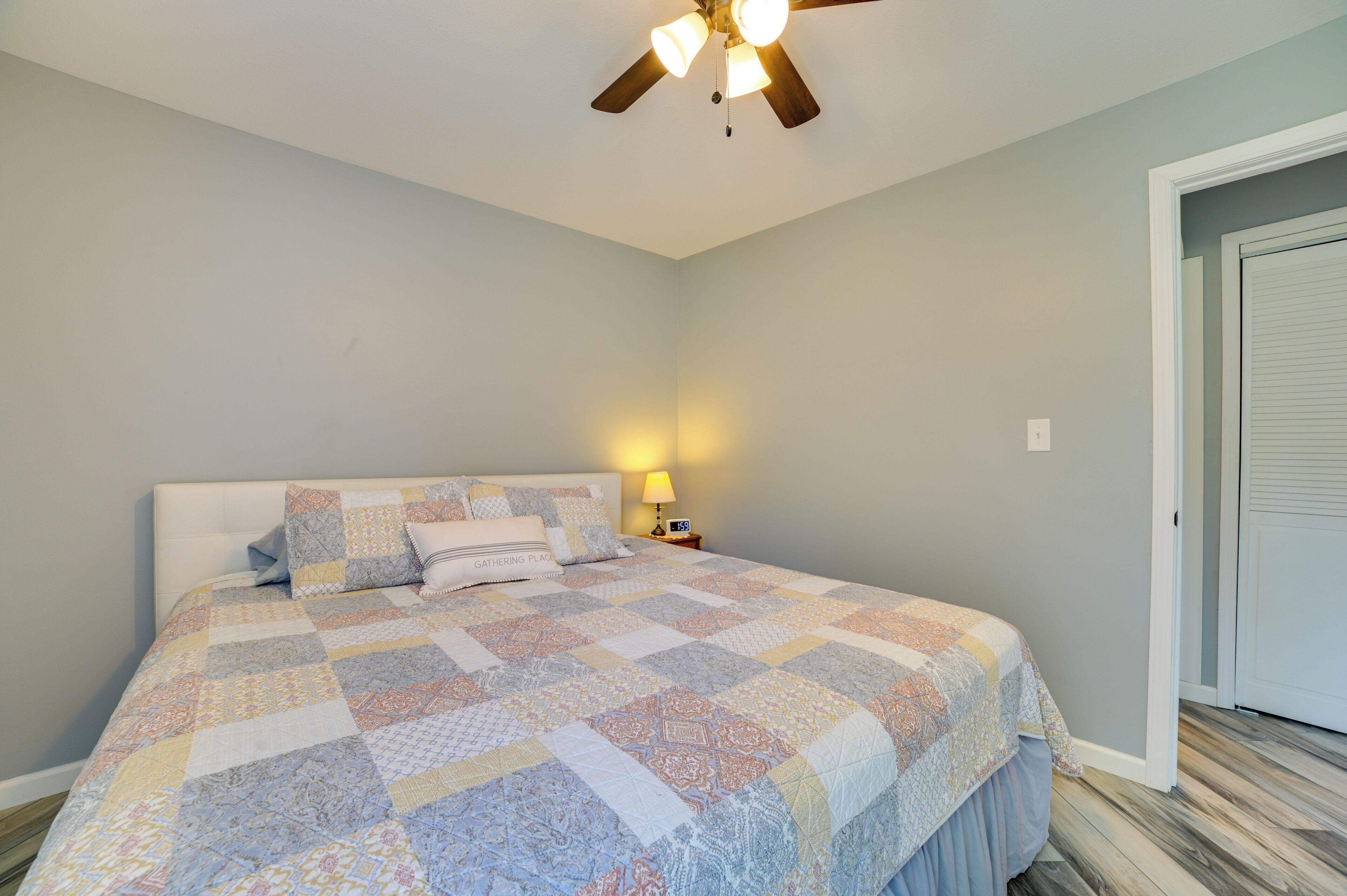A bedroom with a bed, a ceiling fan, a nightstand with a lamp, and a door.