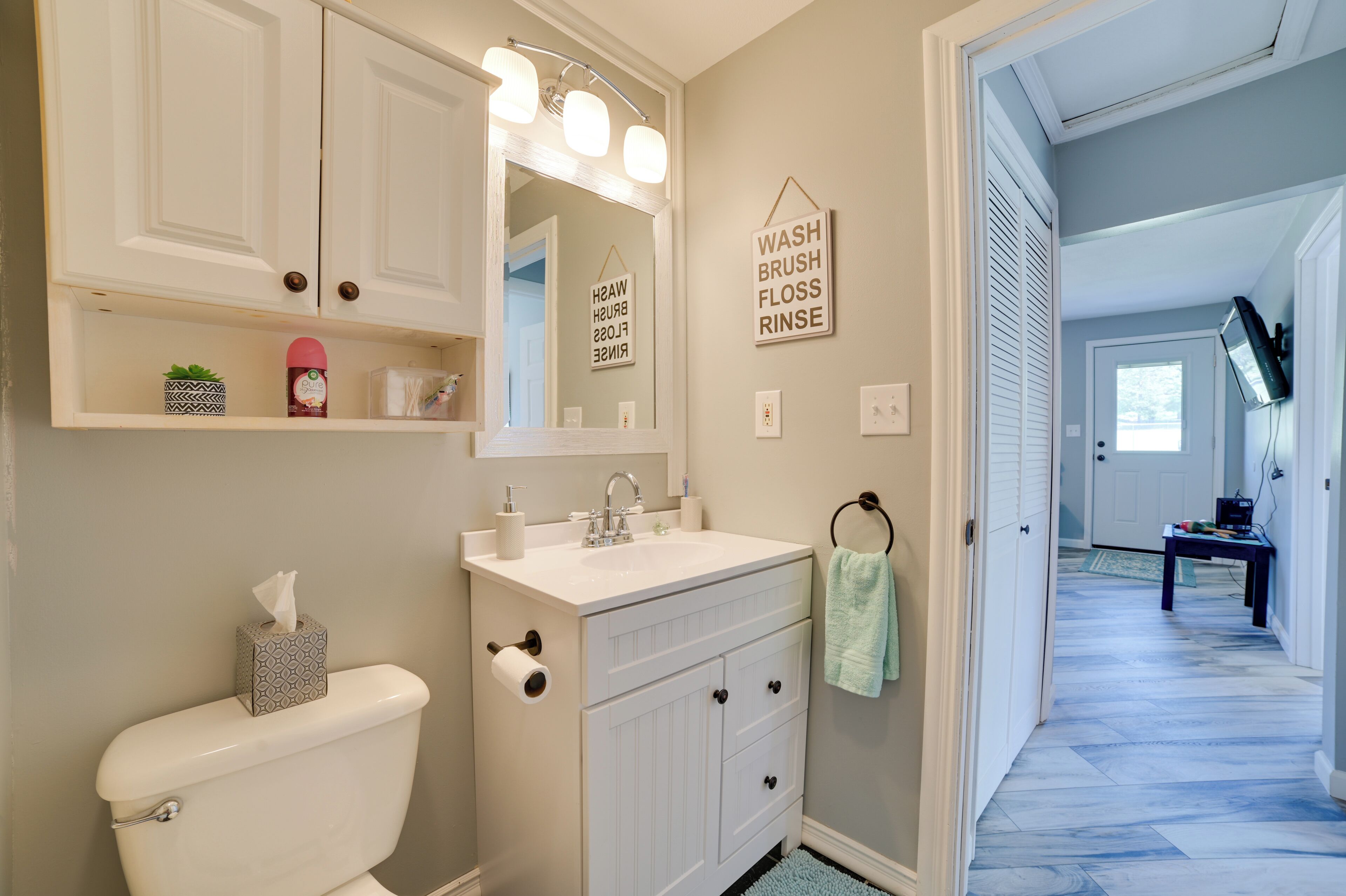 Une salle de bain avec des toilettes, un lavabo et un miroir. Il y a des étagères au-dessus du lavabo où sont rangés des articles de toilette, ainsi qu’une serviette suspendue. Un panneau sur le mur indique : « LAVER, BROSSER, UTILISER DU FIL DENTAIRE, RINCER ».
