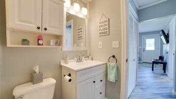 Un baño con inodoro, lavabo y espejo. Hay repisas sobre el lavabo con artículos de tocador y una toalla colgando. Un cartel en la pared dice: 'WASH BRUSH FLOSS RINSE'.