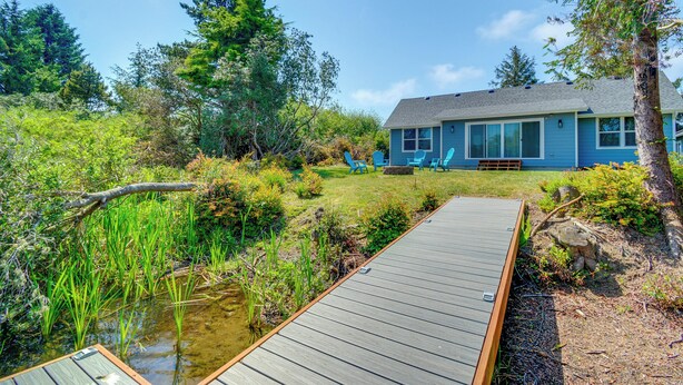 A house with a deck leading to a pond, surrounded by greenery and trees.