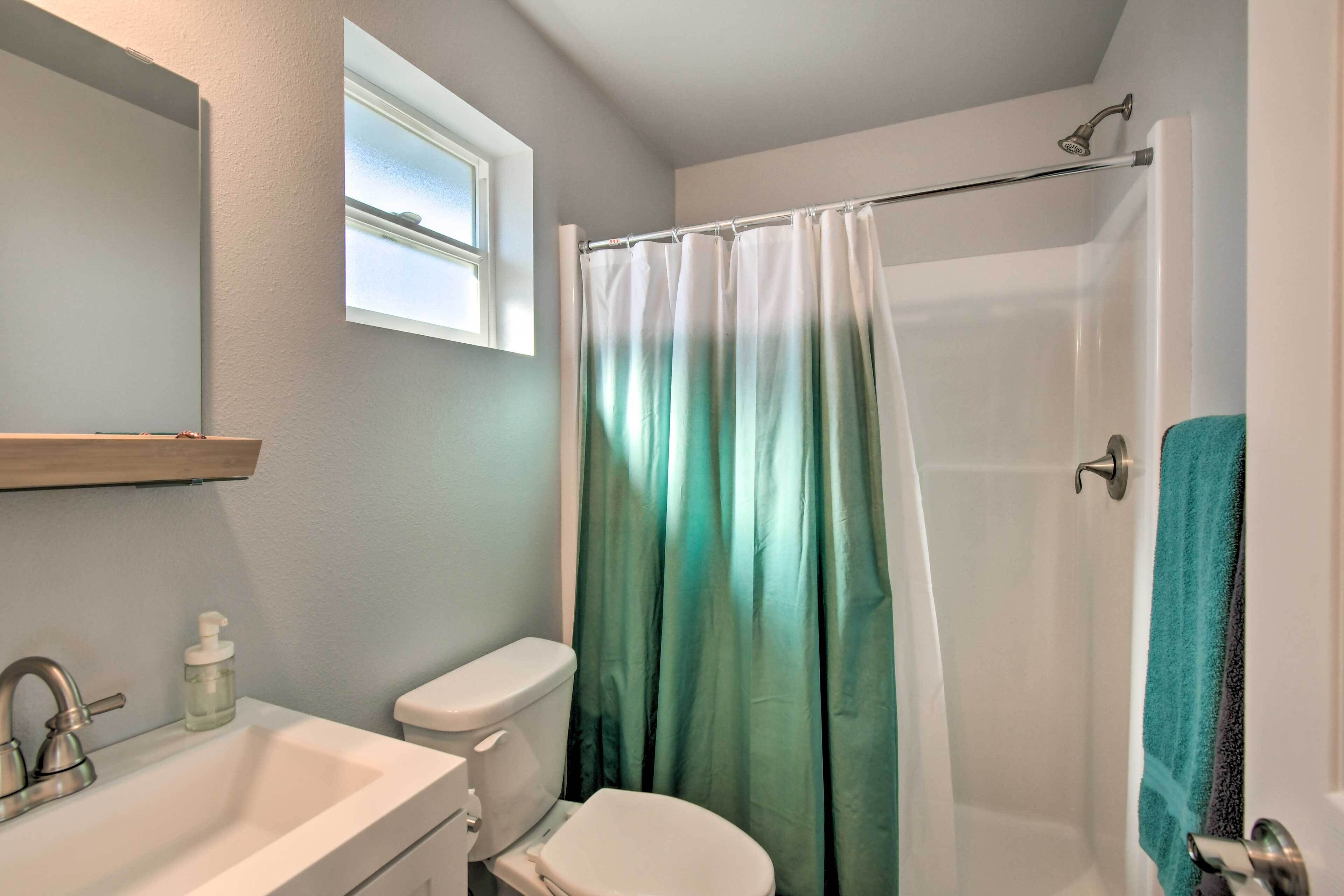 A bathroom with a white sink, a toilet, a shower with a green curtain, and a window above the sink.