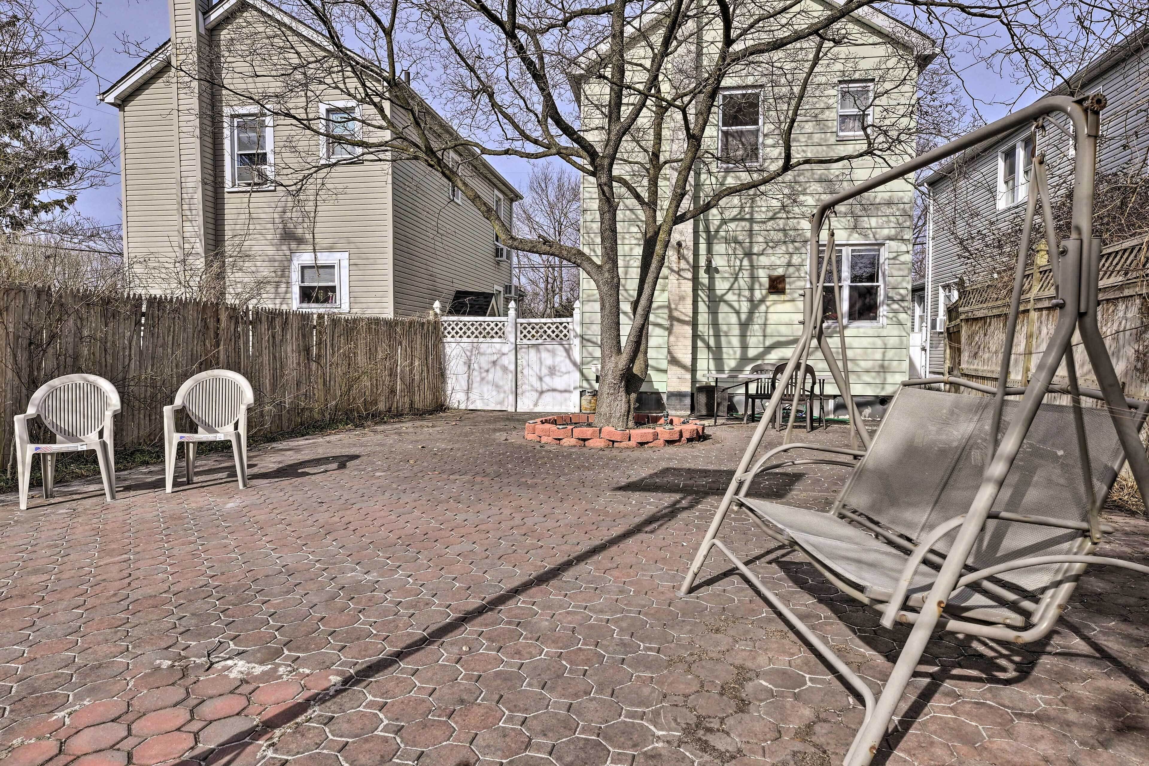 A backyard with a brick patio, two white plastic chairs, a metal swing set, and a wooden fence.