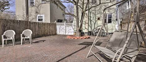 A backyard with a brick patio, two white plastic chairs, a metal swing set, and a wooden fence.