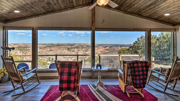 A room with a wooden ceiling, large windows, and a view of a canyon. There are rocking chairs and a table with a rug. - Charming Texas Home w/ Stunning Canyon Views! (Canyon)