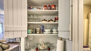 Une cuisine avec des meubles blancs, une table à manger agrémentée d’un chemin de table à motifs floraux, et une variété d’ustensiles de cuisine et de vaisselle rangés sur les étagères.