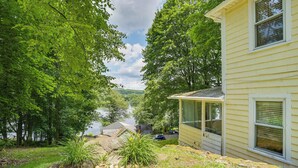 Une maison dotée d’un porche abrité, offrant une vue sur un lac, et d’une cour verdoyante parsemée de rochers et de plantes.