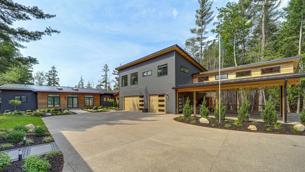 A modern two-story house with a covered driveway, surrounded by a landscaped garden and tall trees. - Clam Lake Vacation Rental w/ Deck & Shared Dock! (Bellaire)