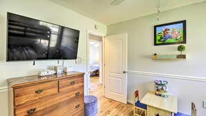Quarto com uma grande TV de tela plana na parede, penteadeira de madeira com gavetas, um banquinho pequeno, uma mesa de madeira com cadeira e uma prateleira com uma pequena planta.