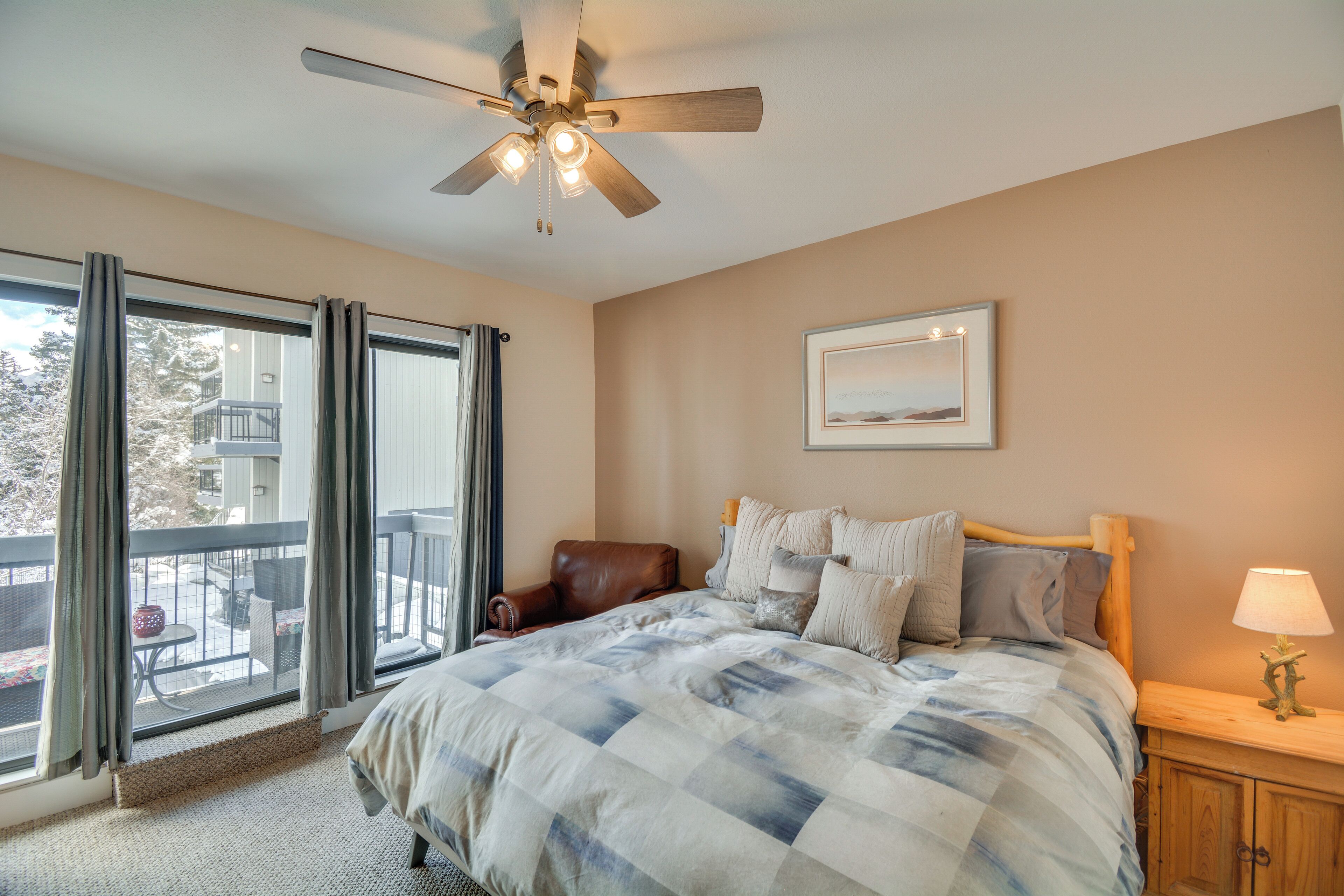 A bedroom with a large bed, a ceiling fan, a nightstand with a lamp, a framed picture on the wall, and a view of the outside through a window.