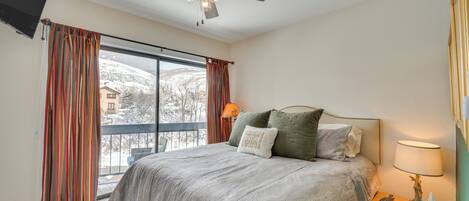 A bedroom with a large bed, a ceiling fan, a nightstand with a lamp, and a view of a snowy landscape through the window.