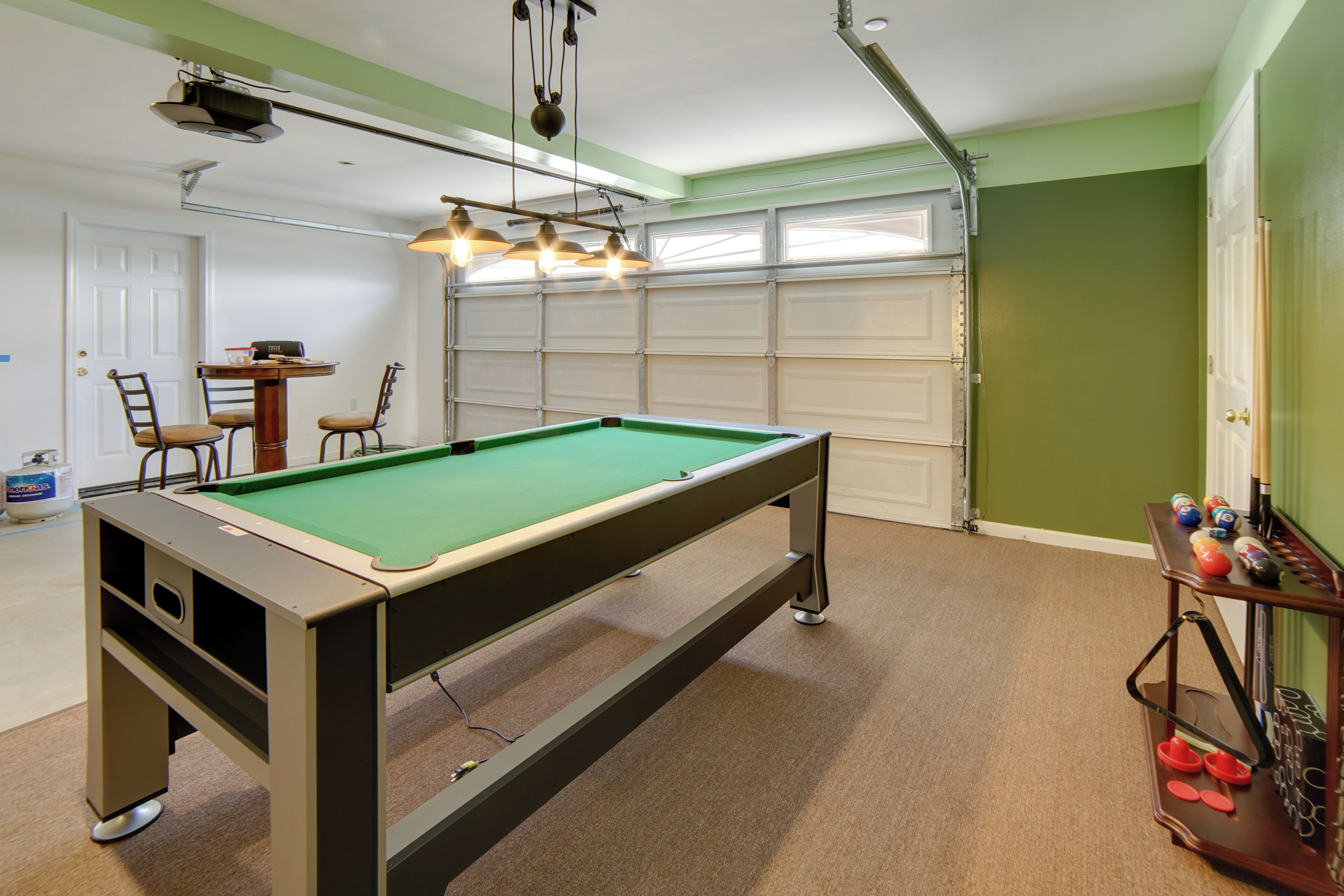 A pool table with chairs, a table with drinks, and a billiards rack in a room with a garage door and green walls.