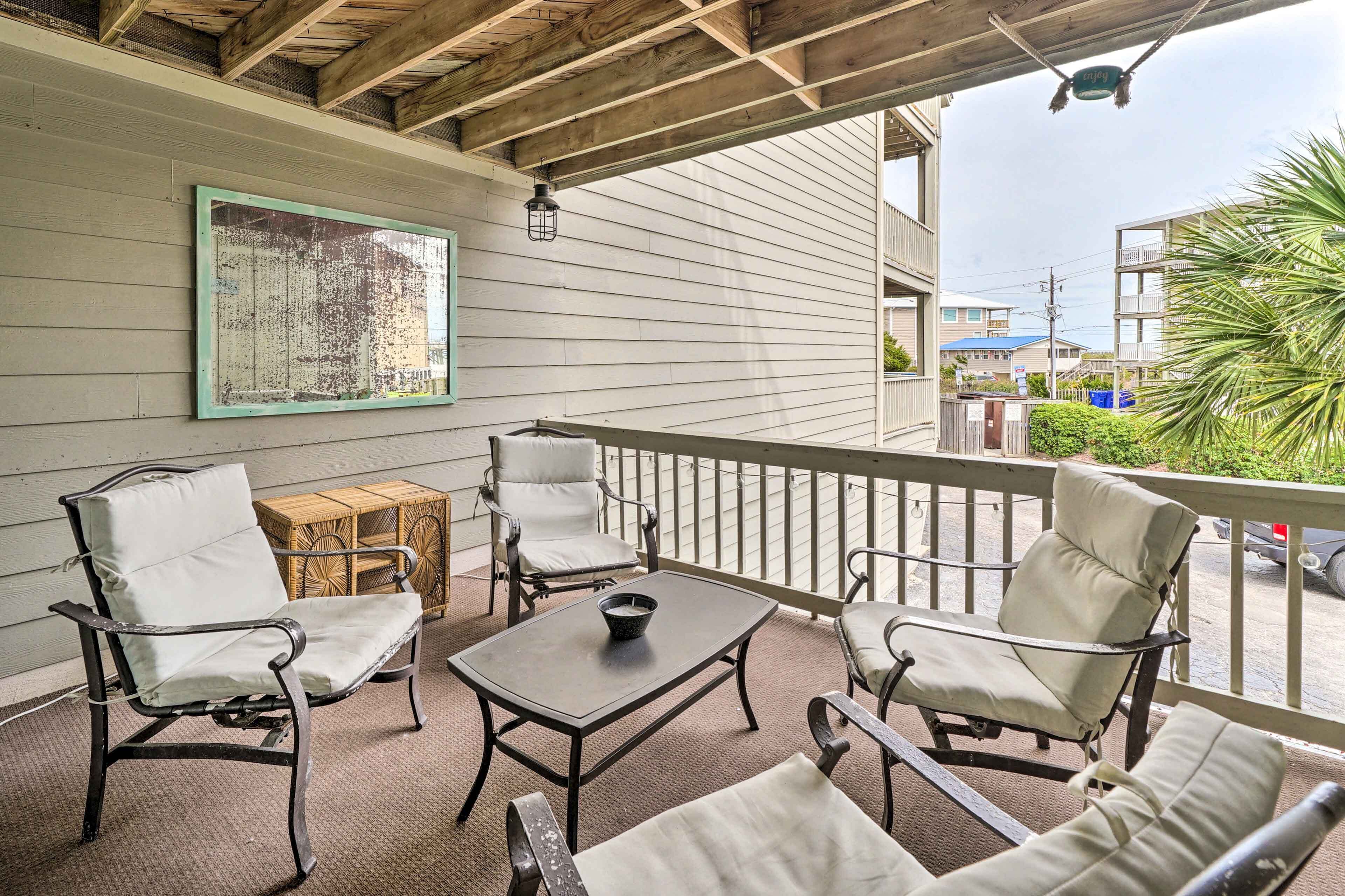 A covered outdoor seating area with metal-framed chairs and a table, a wooden crate, and a framed picture on the wall.