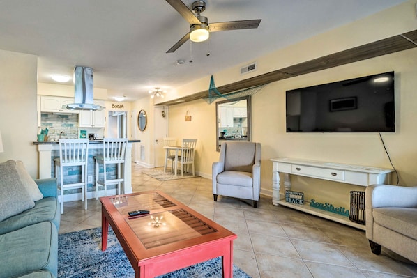 A living room with a TV, a coffee table, and a sofa. There is a kitchen in the background with a dining area.
