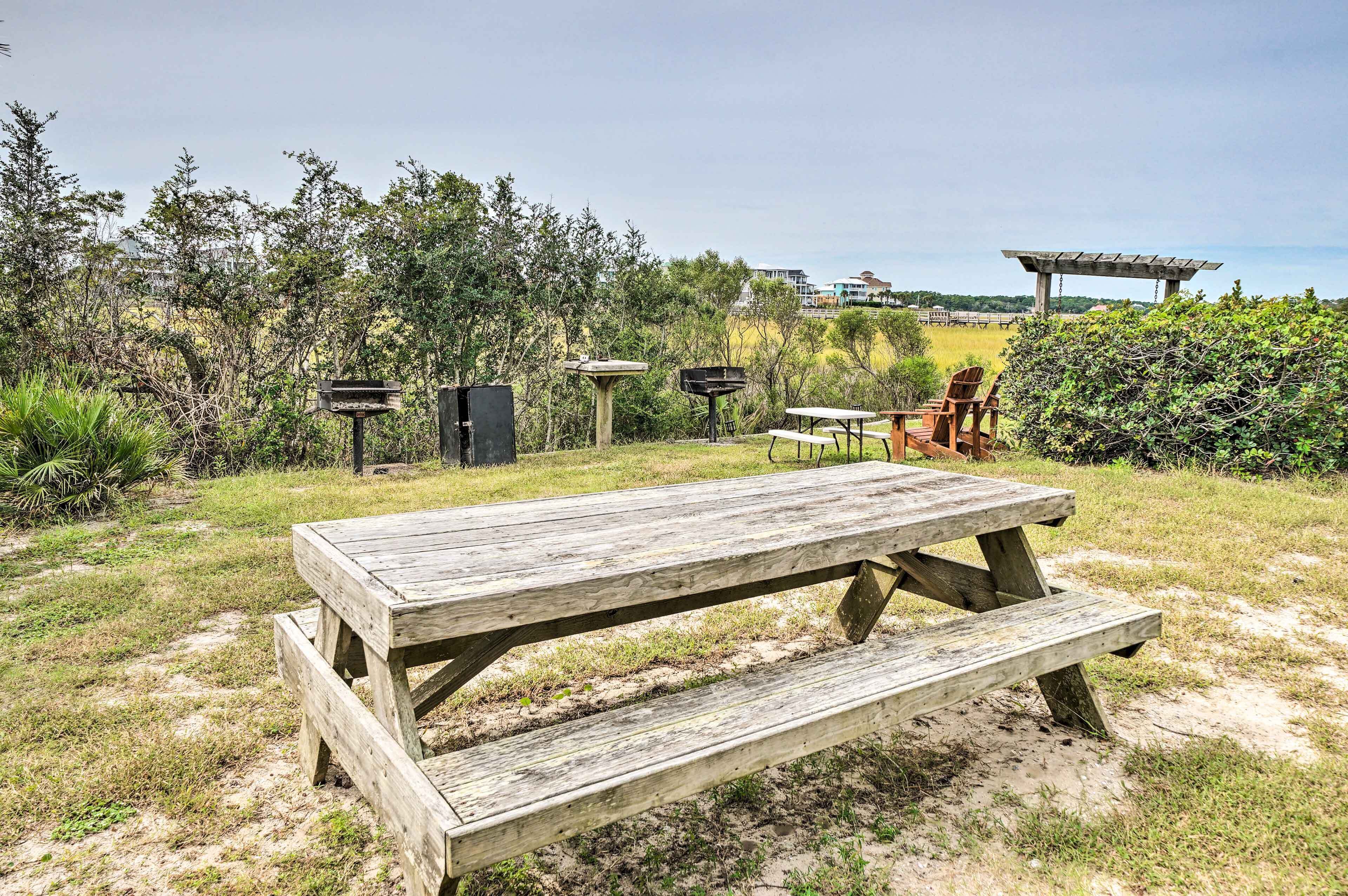 A wooden picnic table with benches in a grassy area with trees and a pergola in the background.