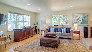 A spacious living room with a large window, a sofa with blue and white pillows, a wooden cabinet, and a decorative rug. - Charming Indianola Home: Walk to Town! (Indianola)