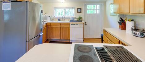 A modern kitchen with wooden cabinets, a stainless steel refrigerator, a dishwasher, and a window with a view of greenery.