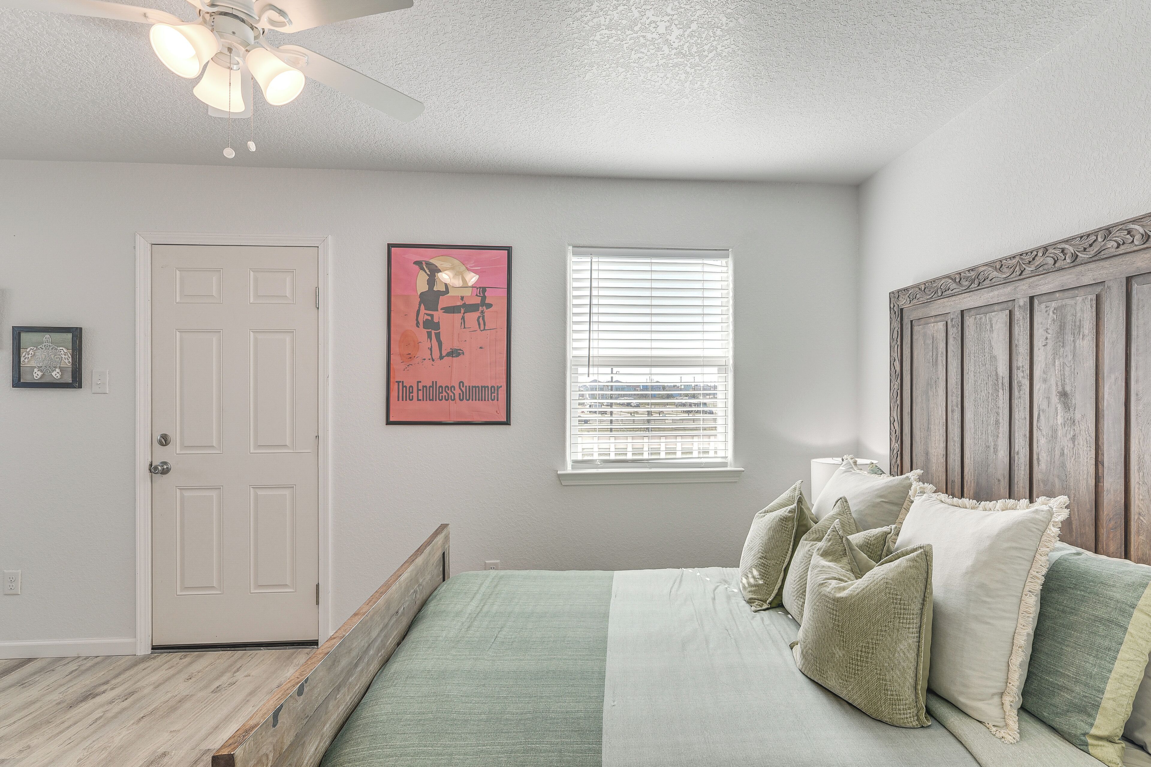 A bedroom with a bed, pillows, a ceiling fan, a door, a window, and a framed picture on the wall.