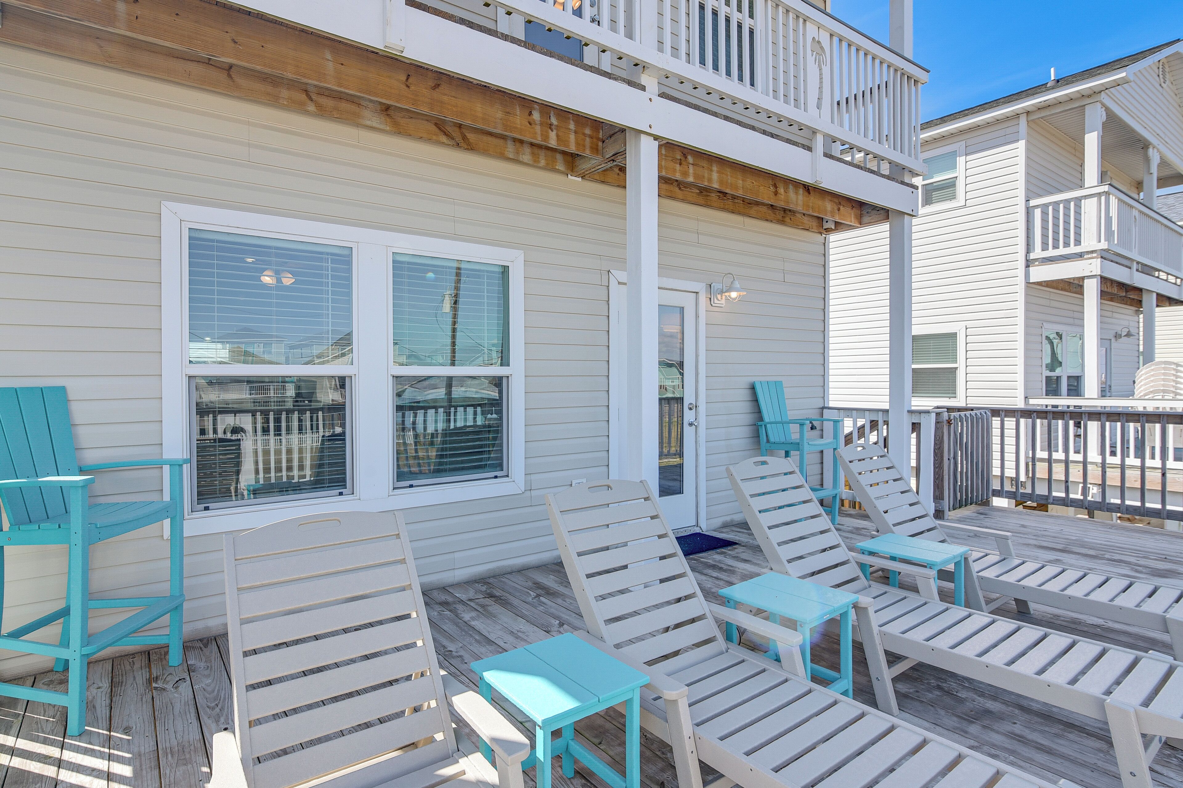 A covered outdoor deck with white and turquoise lounge chairs and tables, wooden railings, and a view of the beach.