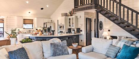 A spacious living room with a high ceiling, a kitchen area with modern appliances, and a staircase leading to an upper level.