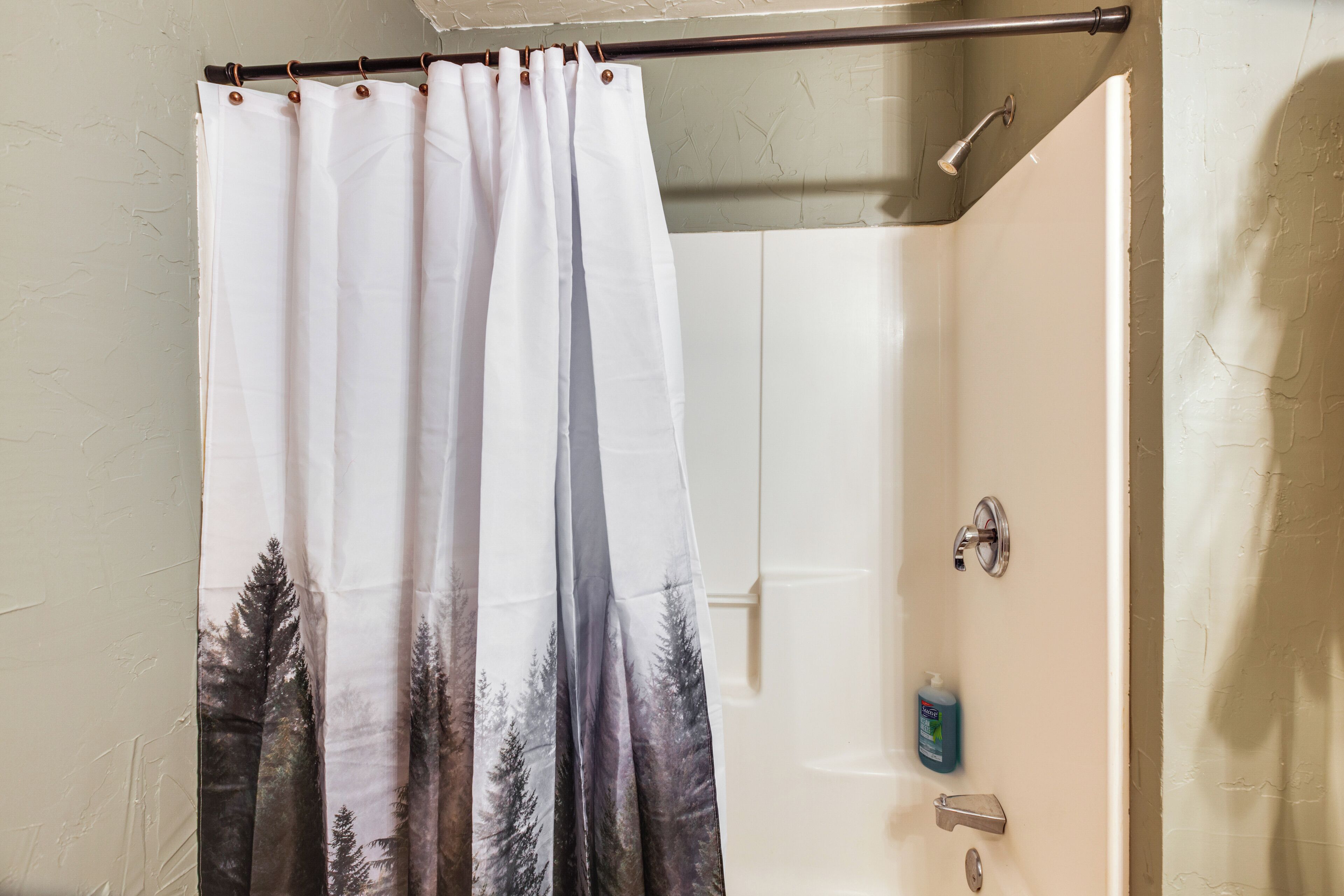 A bathroom with a shower curtain featuring a forest print, a handheld showerhead, and a soap dispenser on the wall.
