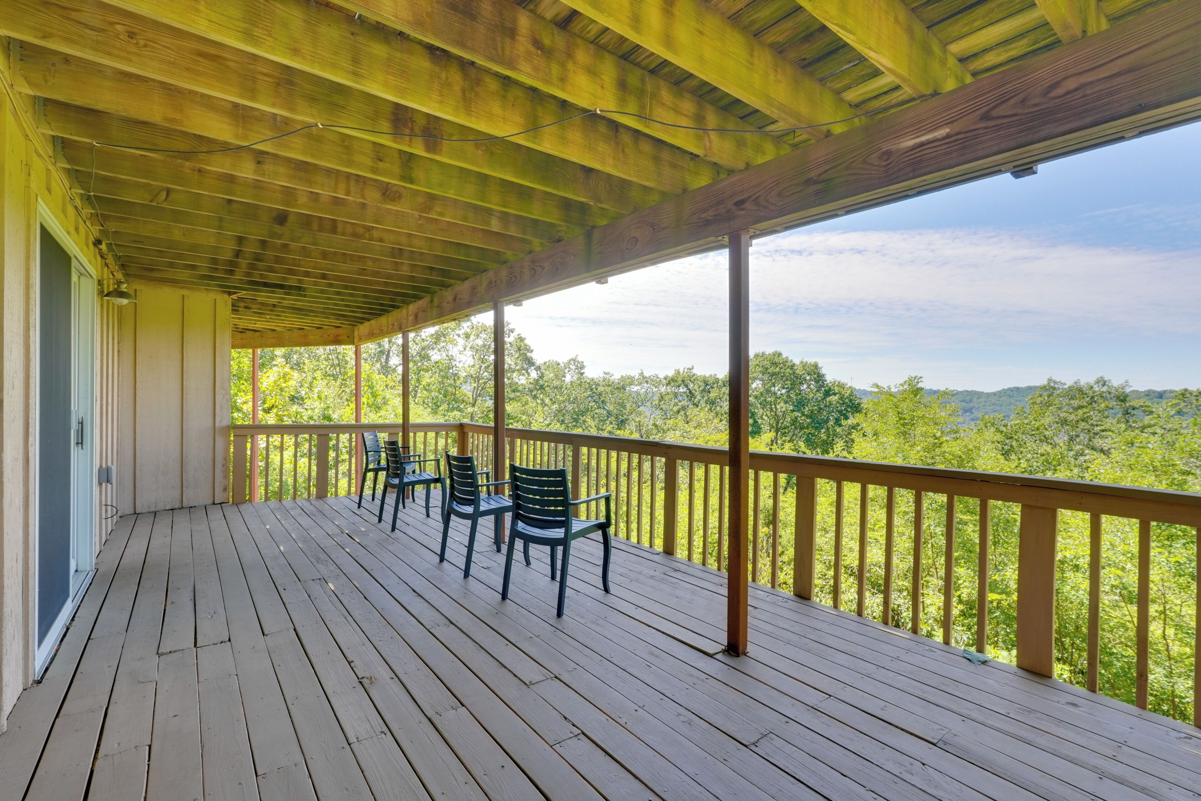 Une véranda couverte avec un plancher en bois et une balustrade, comprenant plusieurs chaises et offrant une vue sur un paysage boisé.