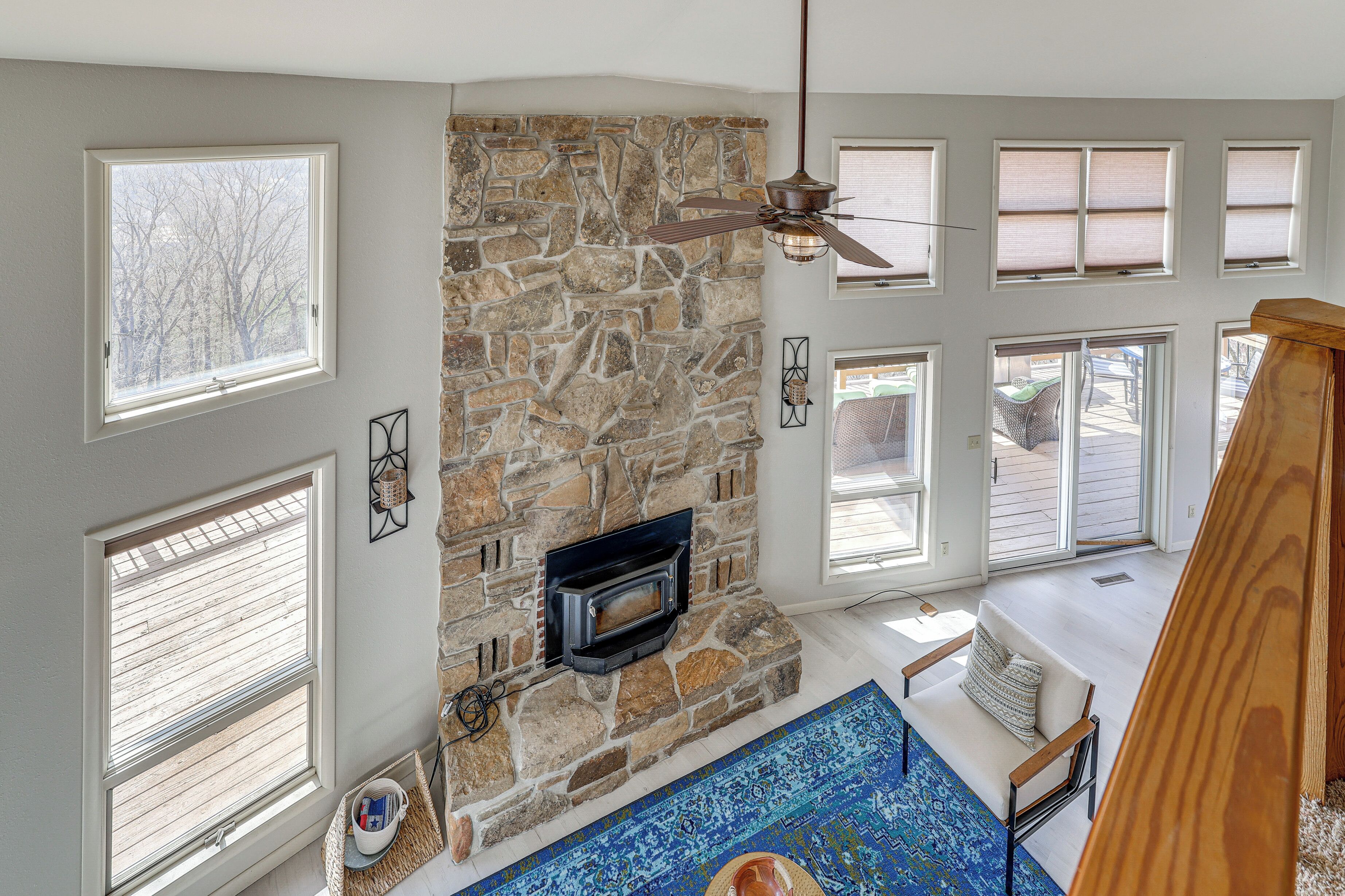 A stone fireplace with a ceiling fan, large windows, and a blue patterned rug.