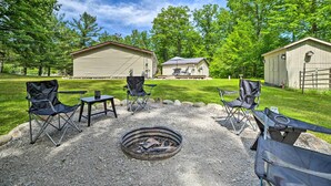 An outdoor seating area with a fire pit, surrounded by four chairs and a table, set against a backdrop of a large building and trees. - Cozy Hale Home Between Long & Loon Lakes! (Hale)