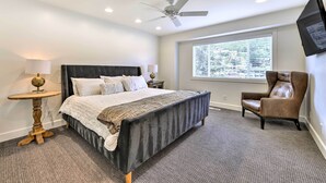 A bedroom with a bed, a chair, a table, a ceiling fan, and a TV mounted on the wall.