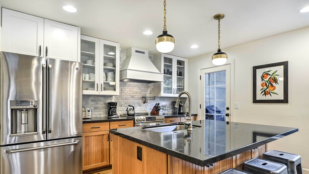 A modern kitchen with stainless steel appliances, wooden cabinets, a large island with stools, and pendant lighting.