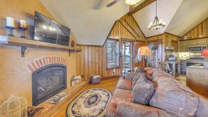 A cozy living room with a fireplace, a large sofa, and a television. - Cozy Ruidoso Cottage w/ Deck - 10 Min to Village! (Ruidoso)