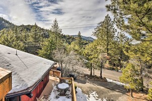 Una terraza de madera con una mesa y sillas cubiertas de nieve, con vista a una carretera y un área boscosa con líneas eléctricas.