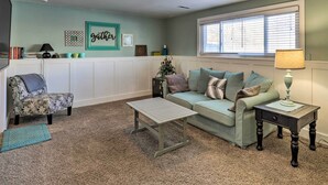 A living room with a sofa, a coffee table, a side table with a lamp, a patterned armchair, a small rug, and a wall with a 'gather' sign.