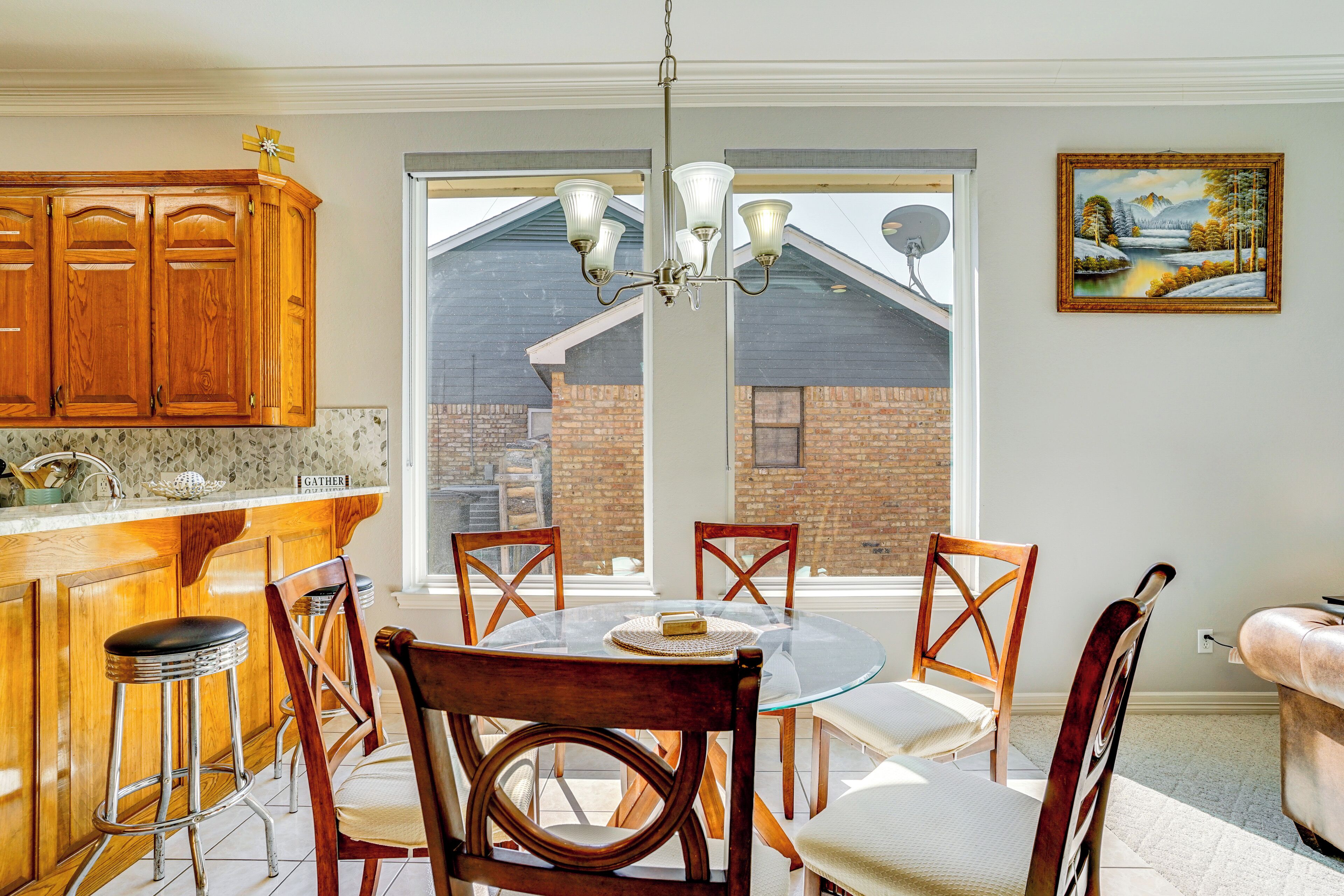 A dining area with a round glass table, wooden chairs, and a chandelier. There is a kitchen area with wooden cabinets and a countertop. A painting is visible on the wall.