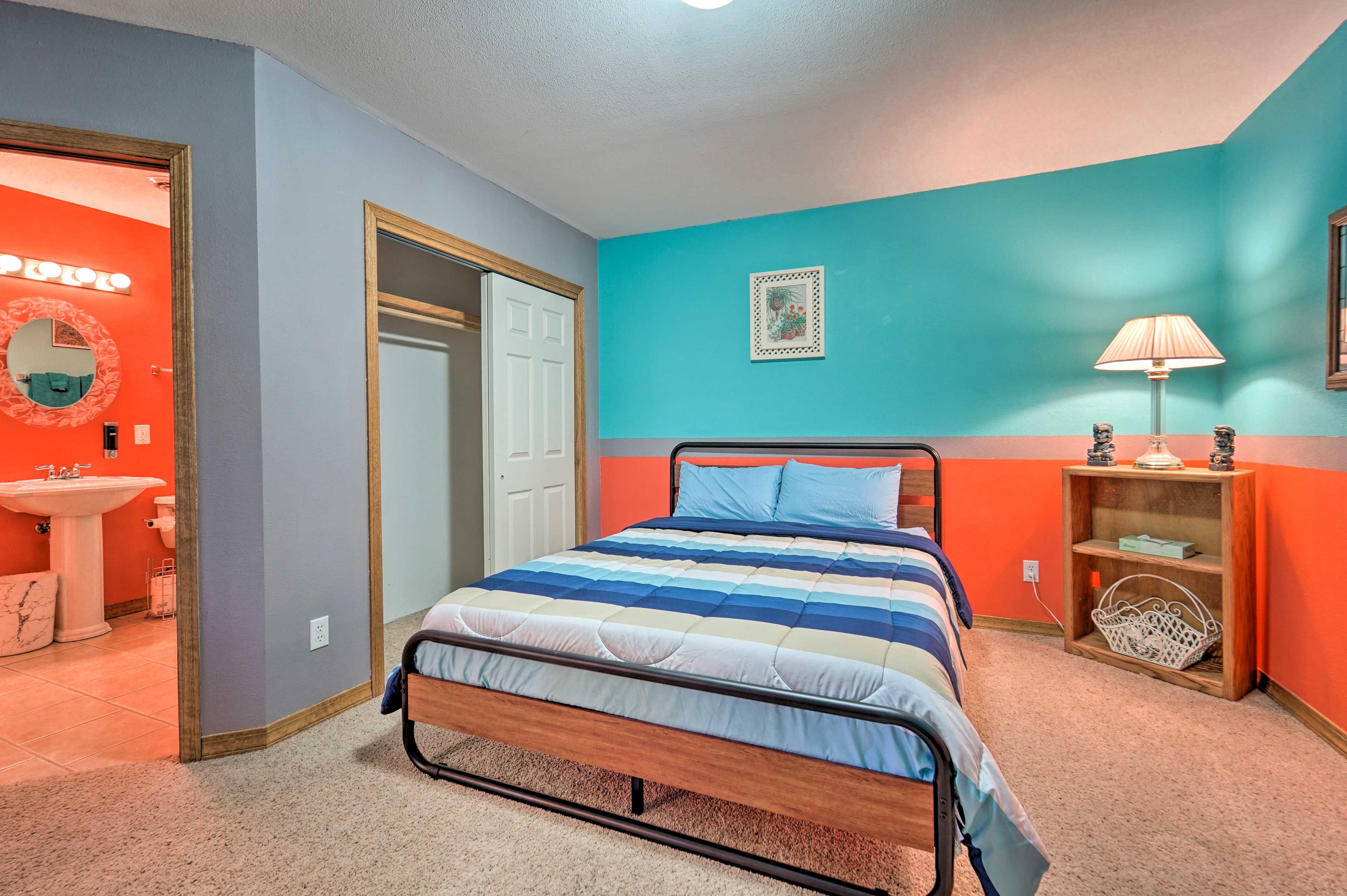 Une chambre à coucher avec un lit, une table de chevet avec une lampe, une étagère avec des objets décoratifs, et une salle de bain visible à travers une porte ouverte.